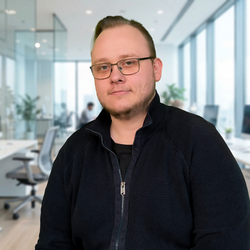 Ein Mann mit kurzen Haaren und Brille trägt eine dunkle, strukturierte Strickjacke über einem schwarzen Shirt und blickt ruhig in die Kamera, während er in einem hellen, modernen Büro steht