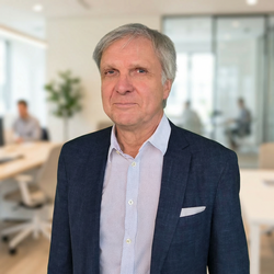 Ein seriös blickender, älterer Mann mit graumeliertem Haar in einem hellblauen Musterhemd und dunkelblauem Sakko steht in einem hellen, modern eingerichteten Büro