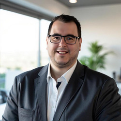 ein freundlich lächelnder Mann mit Brille und kurzem dunklem Haar trägt ein weißes Hemd sowie ein dunkles Sakko und steht in einem hellen, modern gestalteten Innenraum mit einer Pflanze im Hintergrund 