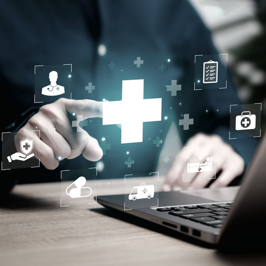 eine Person sitzt an einem Laptop und interagiert mit einer schwebenden, leuchtenden Benutzeroberfläche, die ein zentrales medizinisches Kreuz sowie verschiedene Icons für Gesundheitsdaten und digitale Vernetzung zeigt