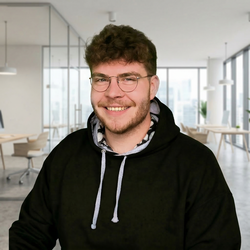 ein lächelnder junger Mann mit braunem Lockenkopf, brille und Dreitagebart trägt einen schwarzen Kapuzenpullover und befindet sich in einem hellen, weitläufigen Großraumbüro