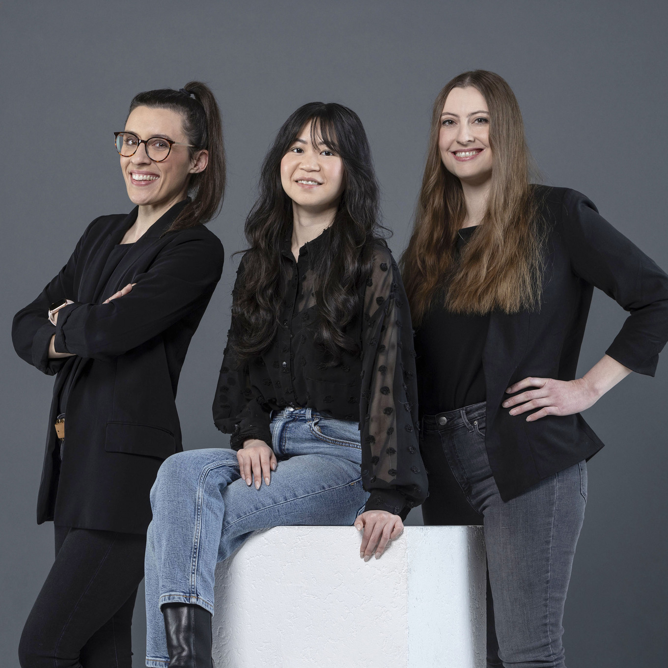 drei lächelnde Frauen posieren vor einem grauen Hintergrund: Die linke Frau mit Brille im schwarzen Blazer hat die Arme verschränkt, die mittlere Frau sitzt auf einem weißen Block und trägt eine schwarze Bluse und Jeans, die rechte Frau steht mit einer hand in der Hüfte