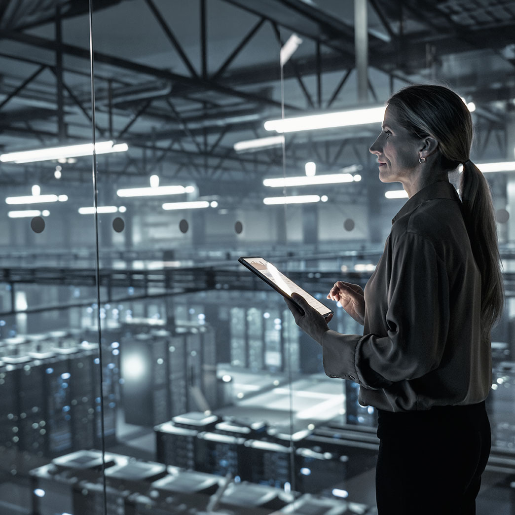 eine junge Frau steht in einem erhöhten Kontrollraum und blickt mit einem Tablet in der Hand über die weitläufigen Server-Reihen eines modernen, bläulich beleuchteten Rechenzentrums