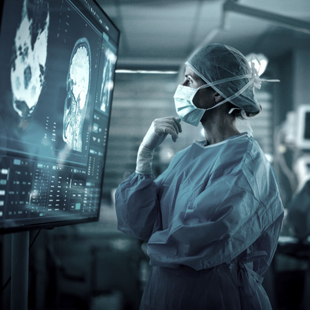 eine chirurgische Fachkraft in voller OP-Montur (Schutzkittel, Haube und Maske) betrachtet in einer klinischen Umgebung konzentriert einen Monitor mit detaillierten medizinischen Bilddaten