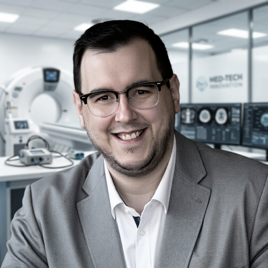 ein freundlich lächelnder Mann mit Brille und kurzem dunklen Haar trägt ein weißes Hemd und graues Sakko vor dem Hintergrund eines medizinischen Behandlungsraums mit einem MRT-Scanner mit einem leicht verdunkelten Hintergrund